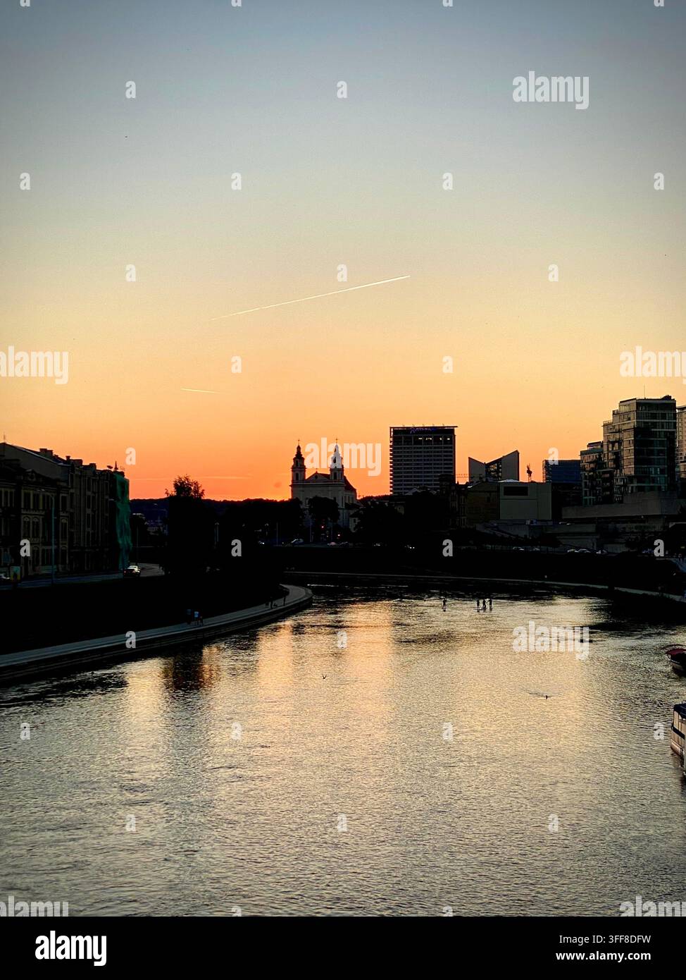 un tramonto dorato sul fiume Neris a Vilnius, Lituania, che mostra le silouette della città, i riflessi dell'acqua e una scia di aereo - Immagine stock catturata con smartphone