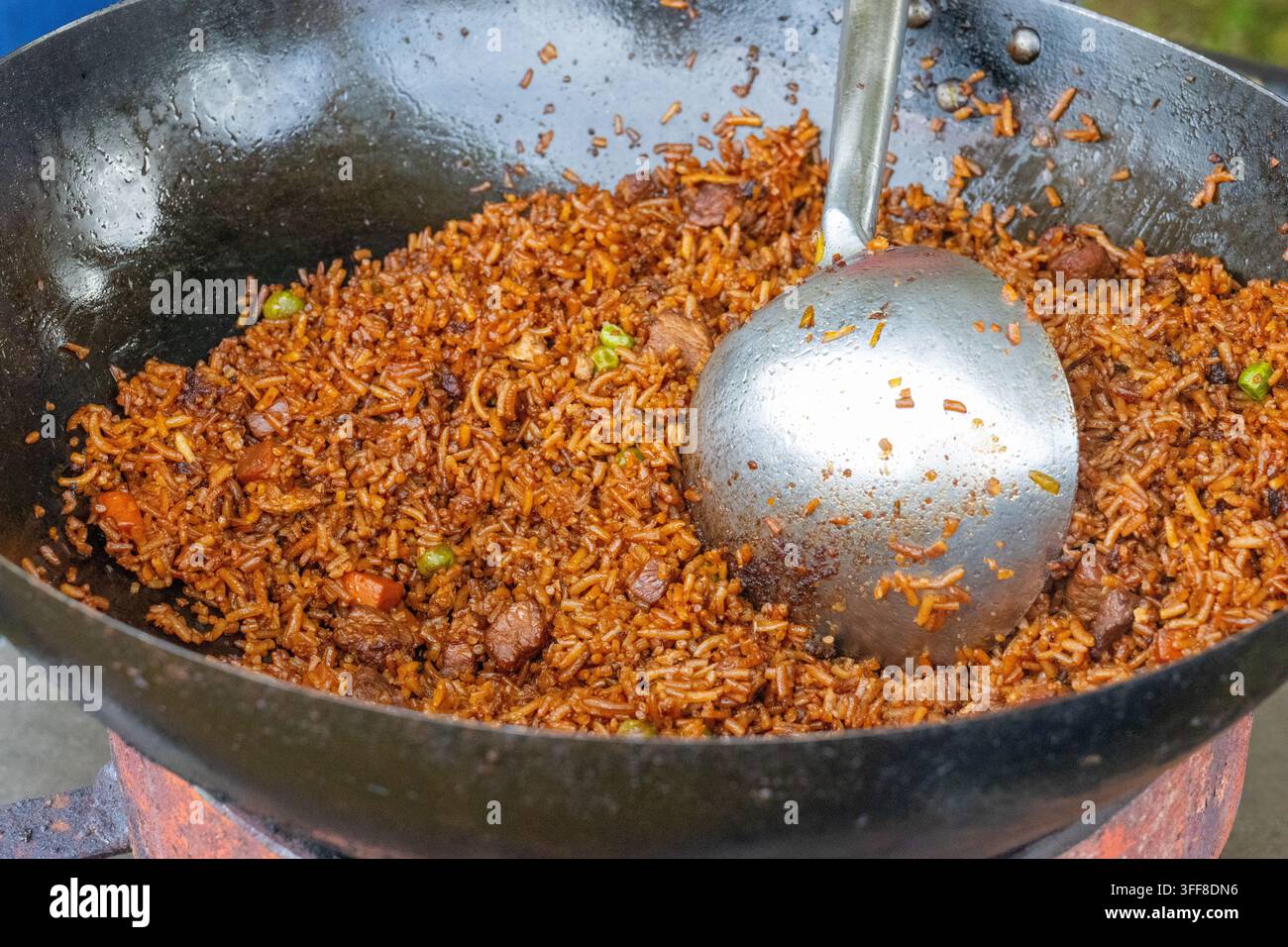 Riso fritto speziato mescolato con carne e piselli cotto in un grande wok nero su un fornello, con un grande mestolo di metallo appoggiato nella padella. Foto Stock