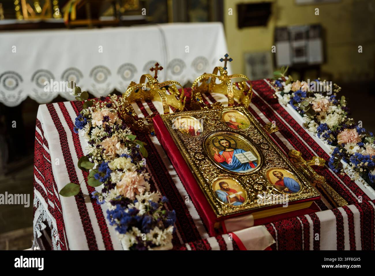 Corone nuziali ortodosse e un libro religioso rivestito d'oro esposti su un panno ricamato con composizioni floreali in un ambiente di luogo di culto Foto Stock