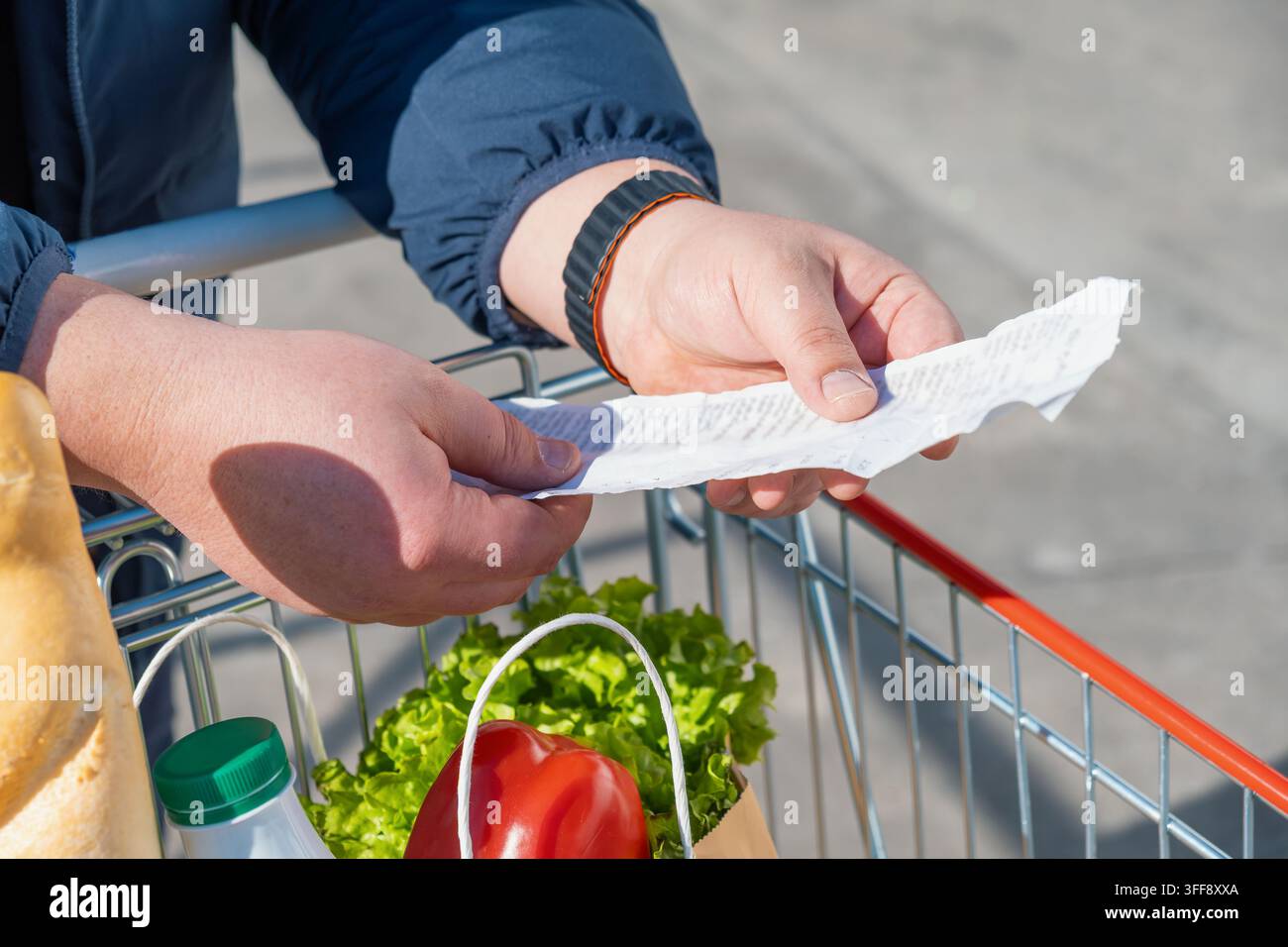 Acquistatore che tiene e controlla una lunga ricevuta del supermercato sopra un carrello con la spesa. Acquisti di generi alimentari quotidiani, aumento dei prezzi dei generi alimentari, inflazione, aumento Foto Stock
