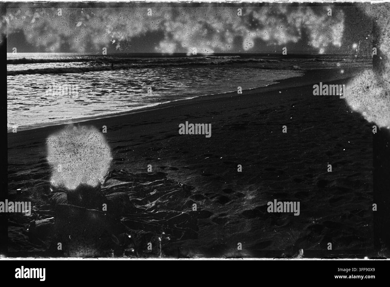 Una spiaggia scura e curva incontra l'acqua increspata in una cornice di pellicola analogica pesantemente distrutta, segnata da perdite di luce, polvere, graffi e bordi ruvidi. Foto Stock