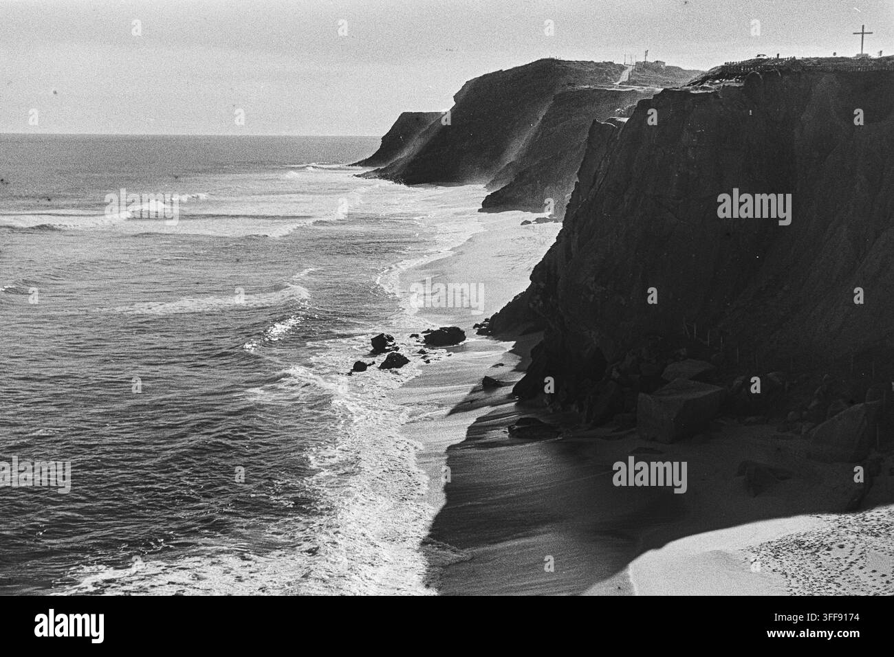 Scogliere marine ripide proiettano ombre profonde su una spiaggia stretta e onde che si infrangono in un paesaggio cinematografico in bianco e nero granuloso. Foto Stock