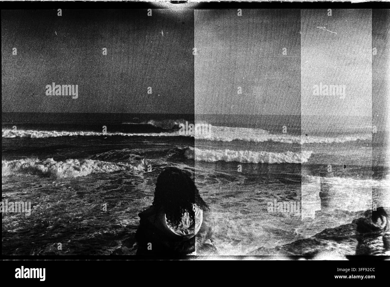 Due figure in controluce affrontano le onde all'interno di esposizioni sulla spiaggia che si sovrappongono su una pellicola bianco e nero fortemente granulosa. Foto Stock