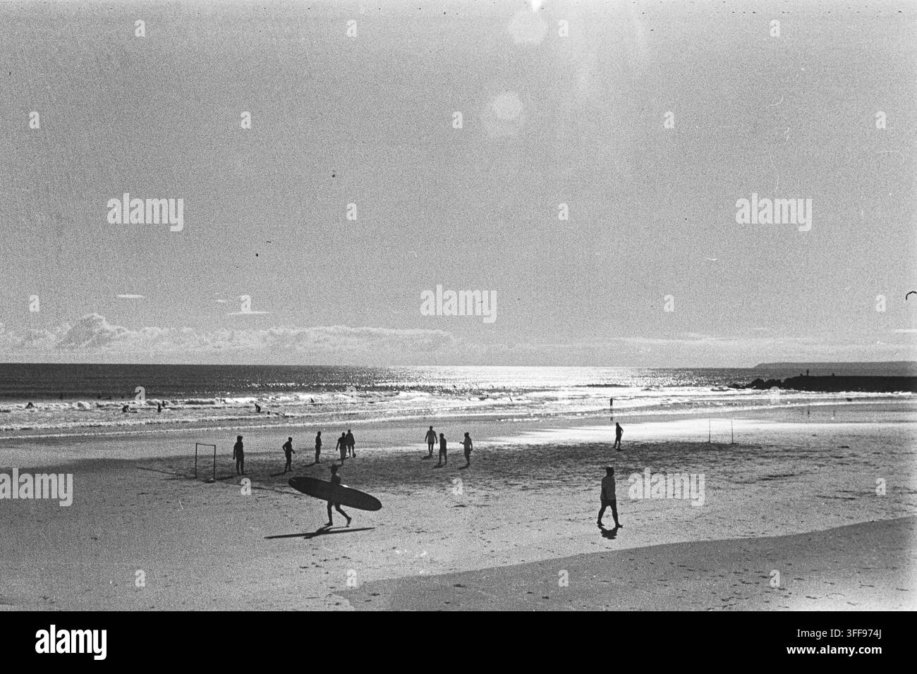 Un surfista trasporta una tavola attraverso una spiaggia ampia mentre piccoli gruppi si radunano vicino all'acqua, catturati in controluce con il grano del film analogico in bianco e nero. Foto Stock