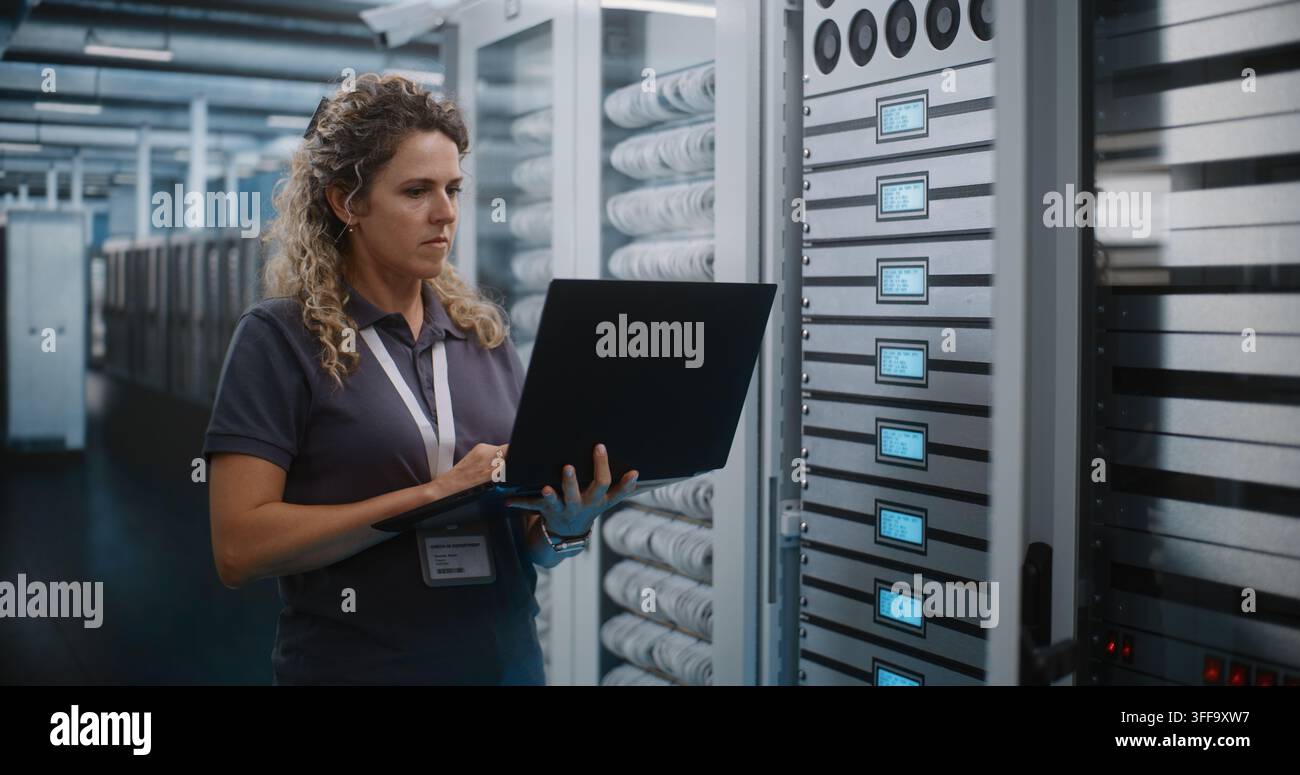 Specialista IT tiene un computer portatile per ispezionare i rack dei server e configurare l’infrastruttura cloud nella sala server del data center. La diagnostica di sistema e la manutenzione dell’hardware garantiscono un uptime di rete senza interruzioni. Foto Stock
