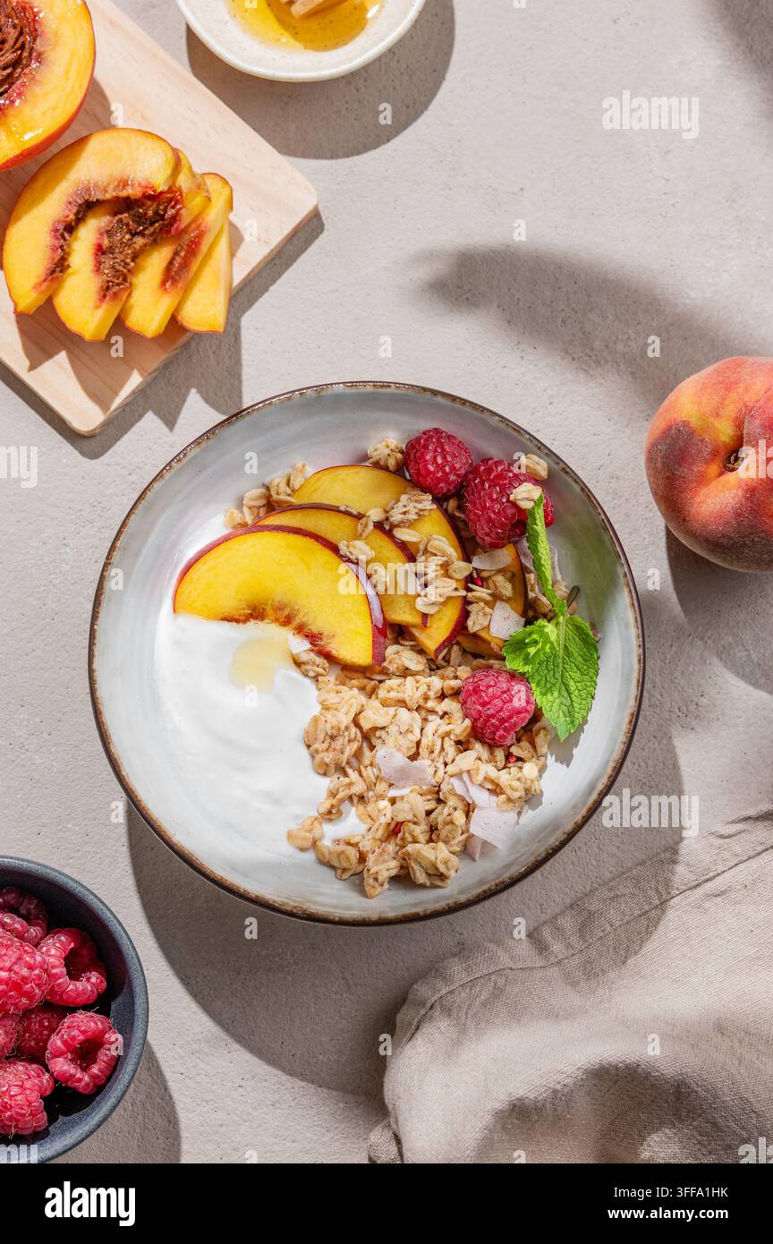 Yogurt greco con granola d'avena o muesli, pesche e lamponi in una ciotola su uno sfondo chiaro con frutta fresca, bacche e ombra mattutina. Sano an Foto Stock