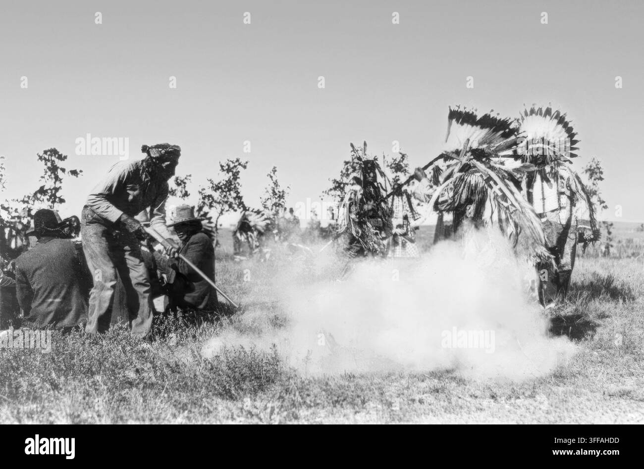 “Danza di guerra”, fotografata da J. A. Anderson intorno al 1911. Un uomo spara verso il suolo, producendo una nuvola di fumo mentre un gruppo di nativi americani con copricapi piumati completi e abiti cerimoniali si trova nelle vicinanze. Una figura fuma una lunga pipa mentre altri osservano l’azione, ambientata su una pianura erbosa aperta. L’immagine riflette le rappresentazioni teatrali della vita rituale indigena dell’inizio del XX secolo, catturando abiti tradizionali, simbolismo del fumo e presenza del gruppo. Foto Stock