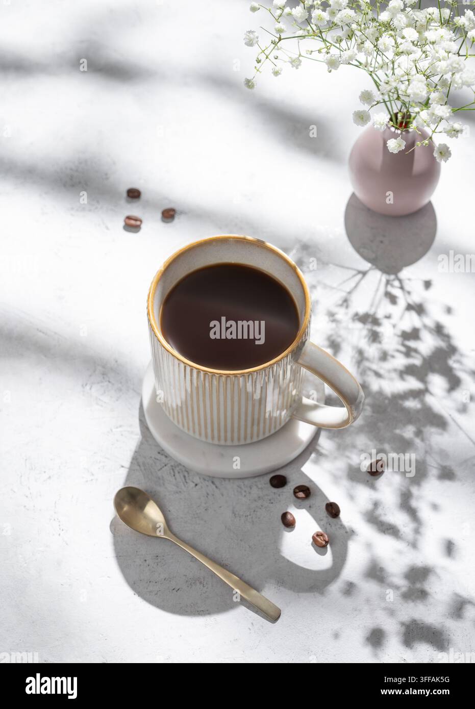 Tazza di caffè appena preparato e mazzo di gypsophila su sfondo leggero e textureggiato con ombre mattutine e chicchi di caffè. Concetto estetico della colazione Foto Stock