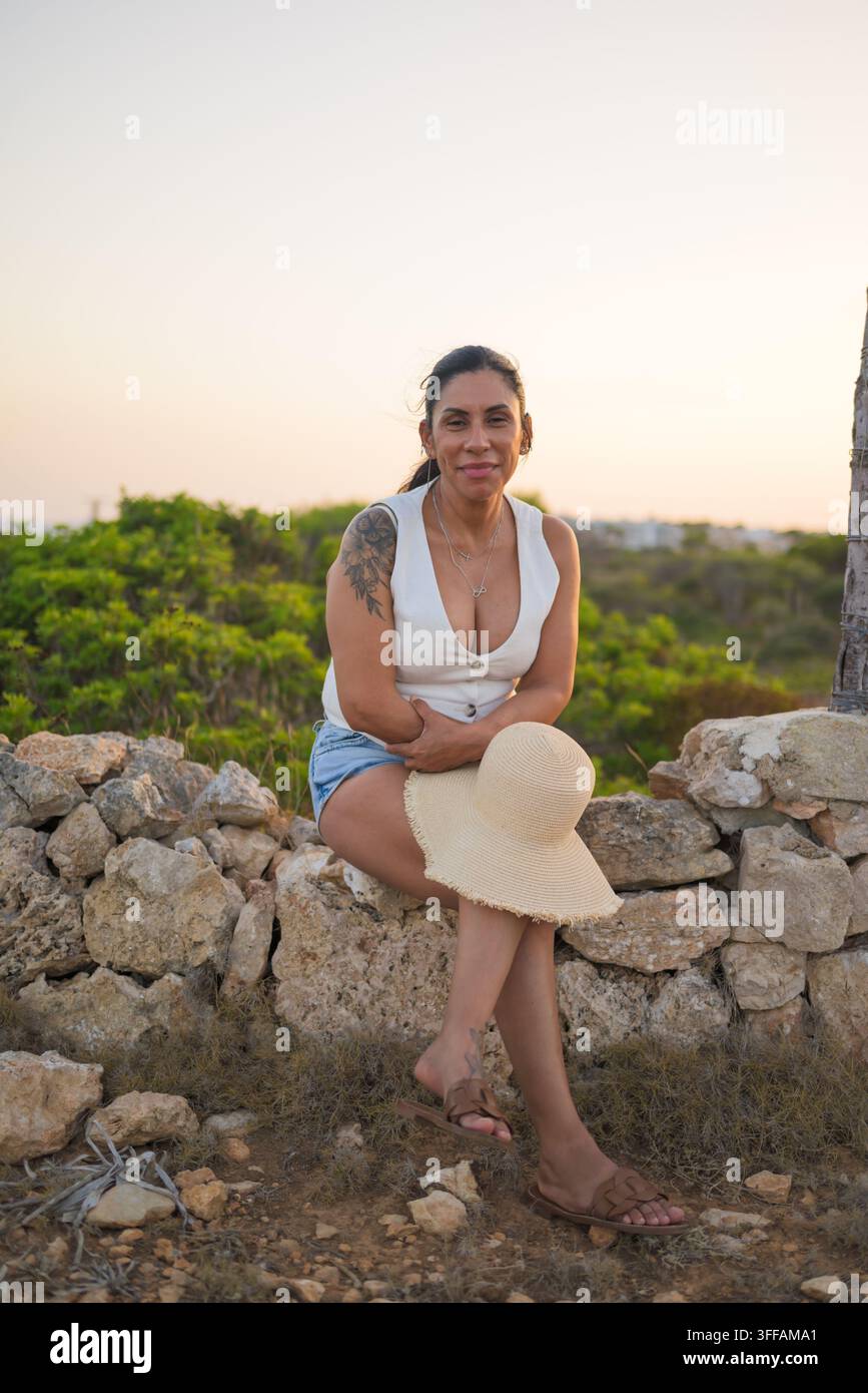 Donna latina sorridente seduta su un muro di pietra durante l'ora dorata Foto Stock