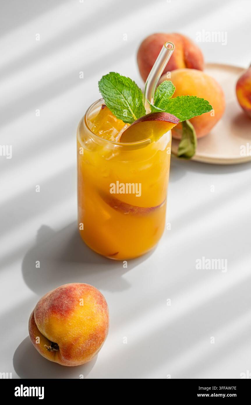Tè freddo alla pesca con menta in un bicchiere su un tavolo bianco vicino alla finestra con frutti freschi e ombra. Concetto di bevanda estiva salutare e rinfrescante per bar o caffè Foto Stock
