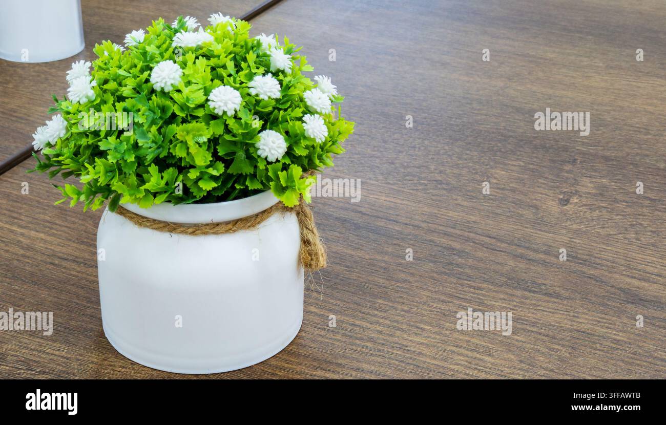 Decorazione da tavolo in un caffè. Pianta verde artificiale con piccoli fiori bianchi in un vaso di ceramica si trova su un tavolo di legno. Composizione centrale accogliente per l'interno. Foto Stock