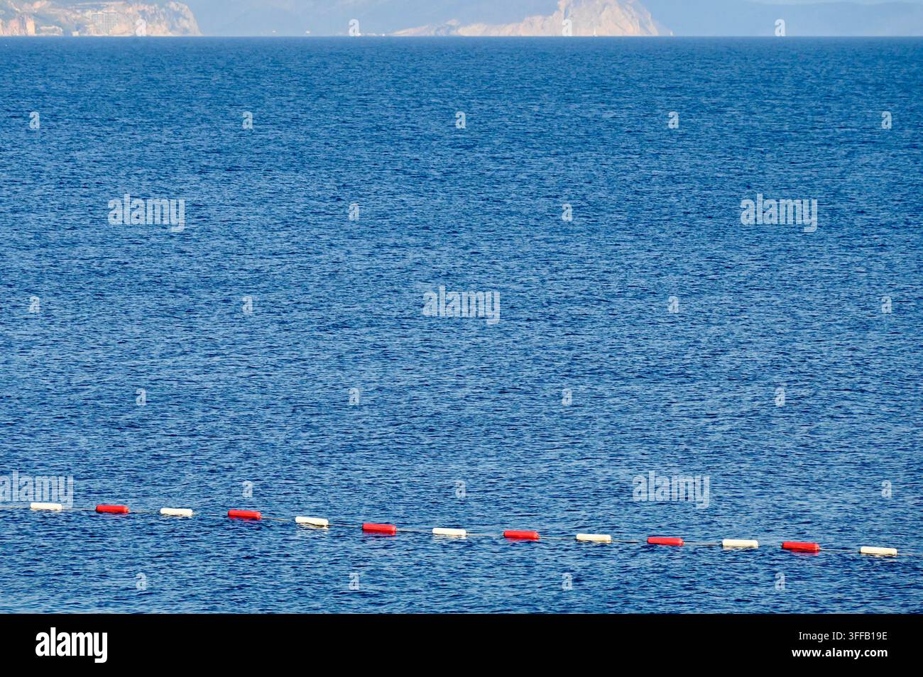 Prima vista di una corda di sicurezza con boe alternate rosse e bianche in plastica che segnalano un'area di nuoto nel mare blu profondo, con una costa sfocata sullo sfondo Foto Stock