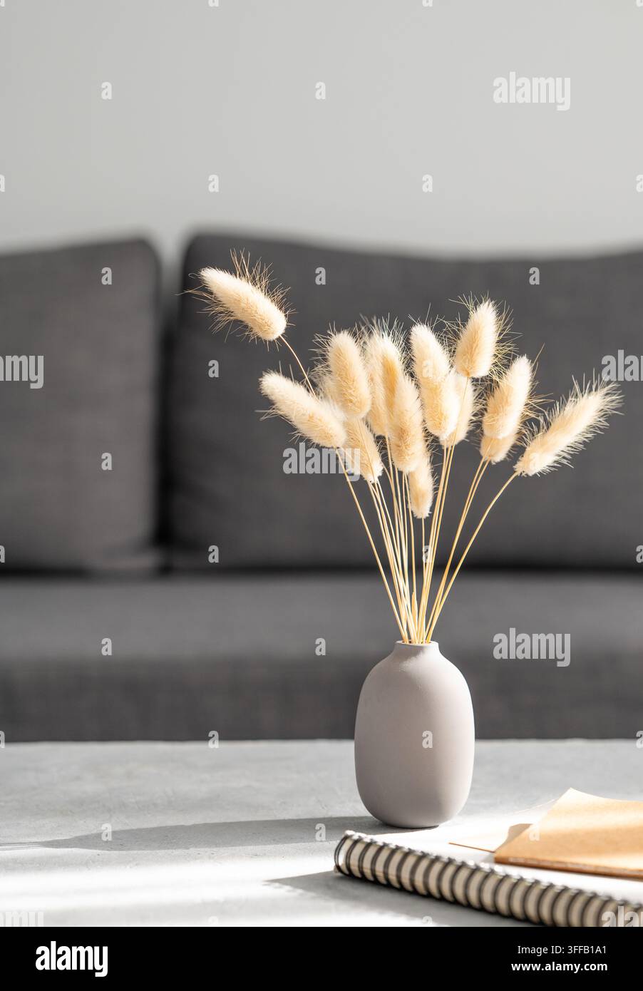 Vaso con erba secca a coda di coniglio e taccuino su tavolo grigio davanti a un divano in stoffa scura. Concetto di decorazione domestica minimalista. Vista frontale, spazio per il testo. Foto Stock