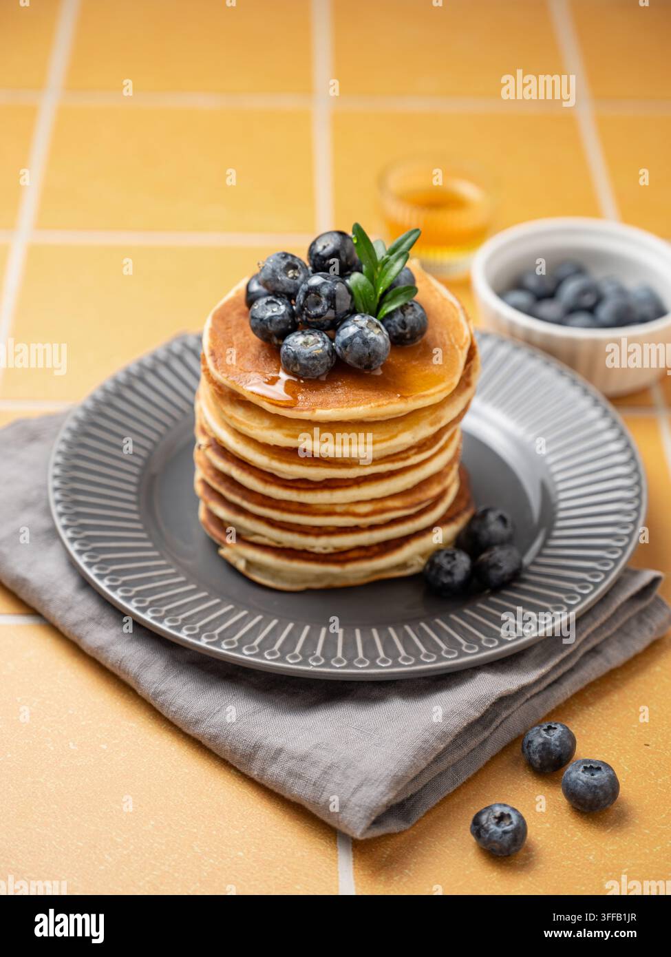 Pancake con mirtillo su un piatto grigio su uno sfondo di piastrelle gialle con frutti di bosco freschi, miele e tovagliolo. Un concetto per colazione o brunch per un menù di caffè Foto Stock