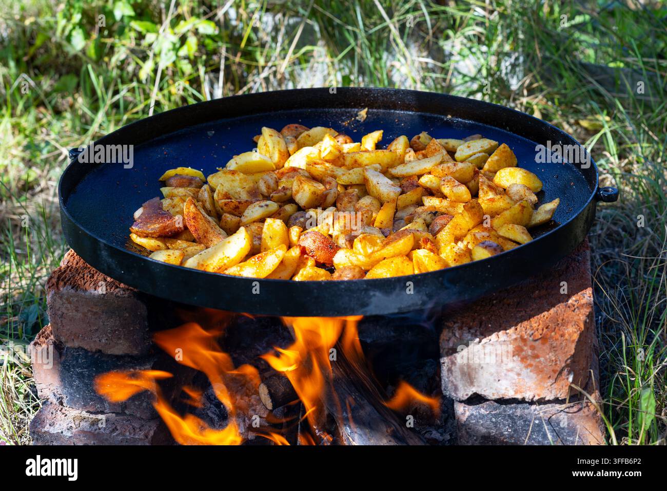 Patate condite che cuociono in padella su fuoco aperto. Wedges di patate condite che cuociono in una grande padella su un fuoco di legna aperto all'aperto. Foto Stock