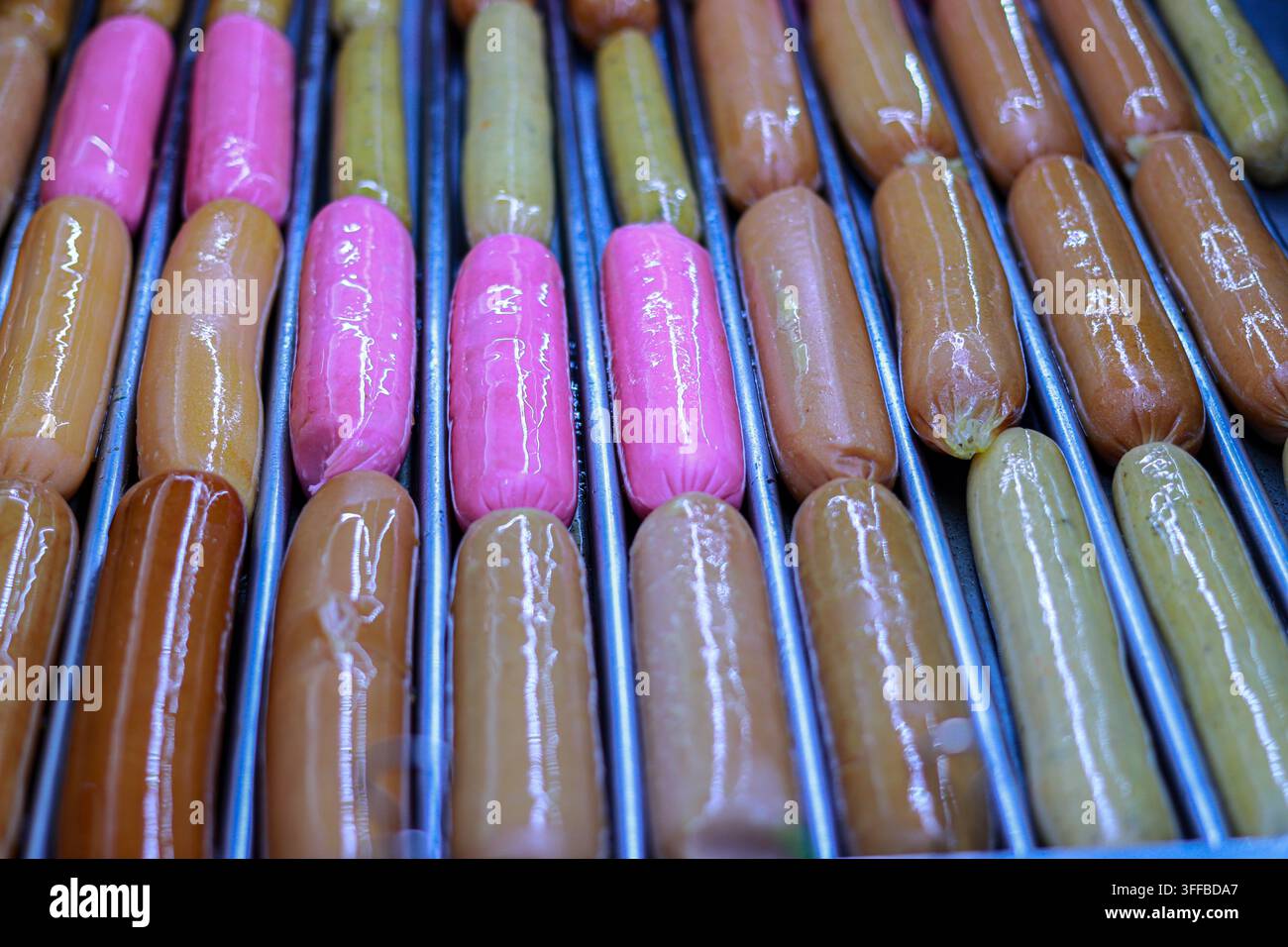 Primo piano di salsicce e hot dog multicolori su una griglia a rulli in metallo, che formano un motivo a griglia ripetitivo e colorato. Foto Stock