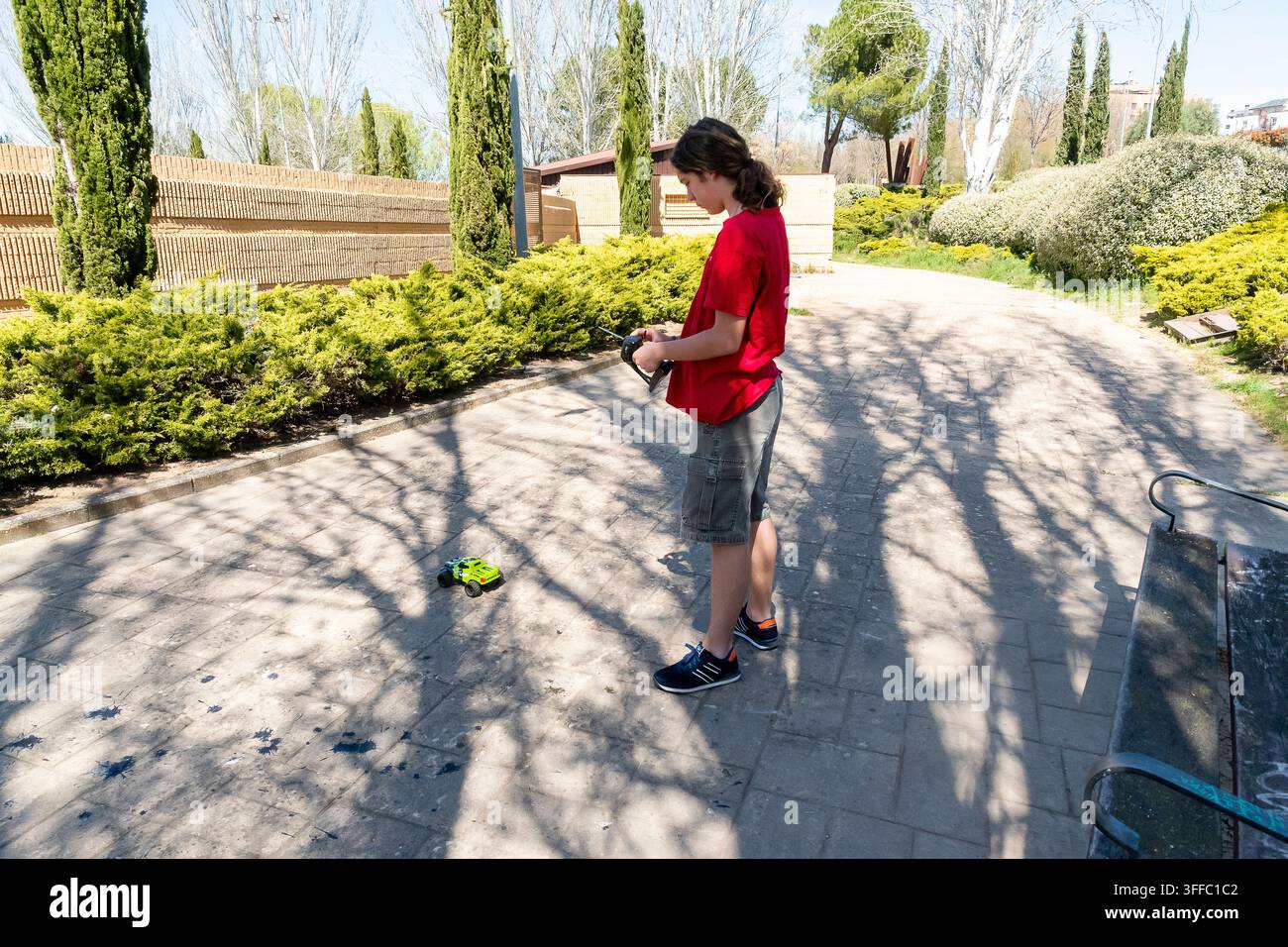 Un ragazzo si trova in un parco, concentrato su una macchinina giocattolo a terra, mentre gli alberi proiettano ombre intorno a lui. Foto Stock