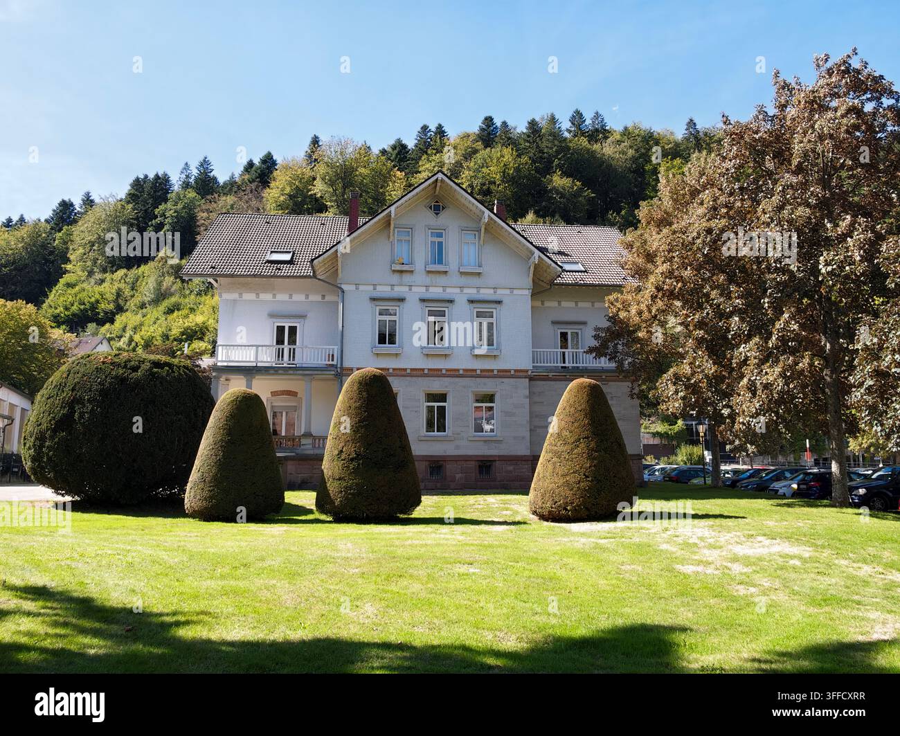 La villa bianca di Bad Herrenalb si erge accanto ad arbusti ben curati e alberi alti su un terreno soleggiato, con una collina boscosa sullo sfondo, il cielo blu sopra e p Foto Stock
