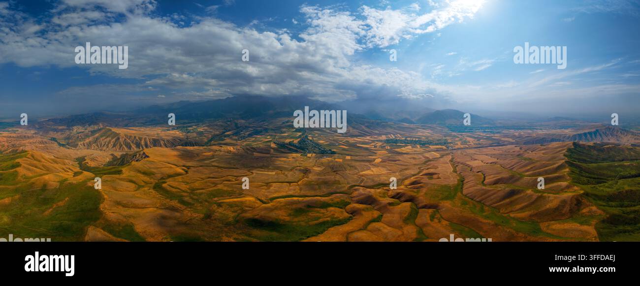 Ampia vista aerea panoramica cattura colline aspre in primo piano che si trasformano in pianure agricole e montagne lontane nel Kirghizistan. Un cielo drammatico presenta un blu brillante e nuvole sparse, con la luce del sole che illumina il paesaggio. Foto Stock