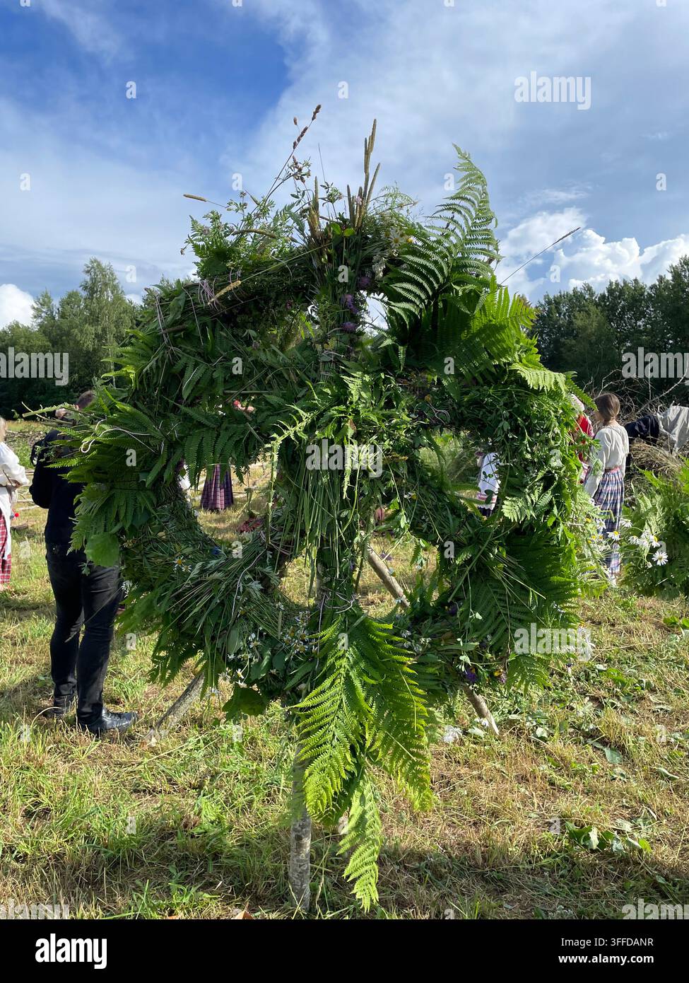Un simbolo tradizionale realizzato con frondi di felce verde lussureggianti, erbe di campo e fiori selvatici installato all'aperto in un prato soleggiato durante il solstizio d'estate - Immagine stock catturata con smartphone