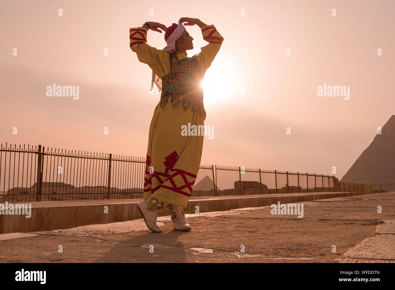 Ballerina folk egiziana in galabiya ricamata con ornamenti esegue una posa trionfale delle braccia, illuminata di spalle dal sole fiammeggiante nell'alone sulla terrazza di pietra delle piramidi di Giza Foto Stock