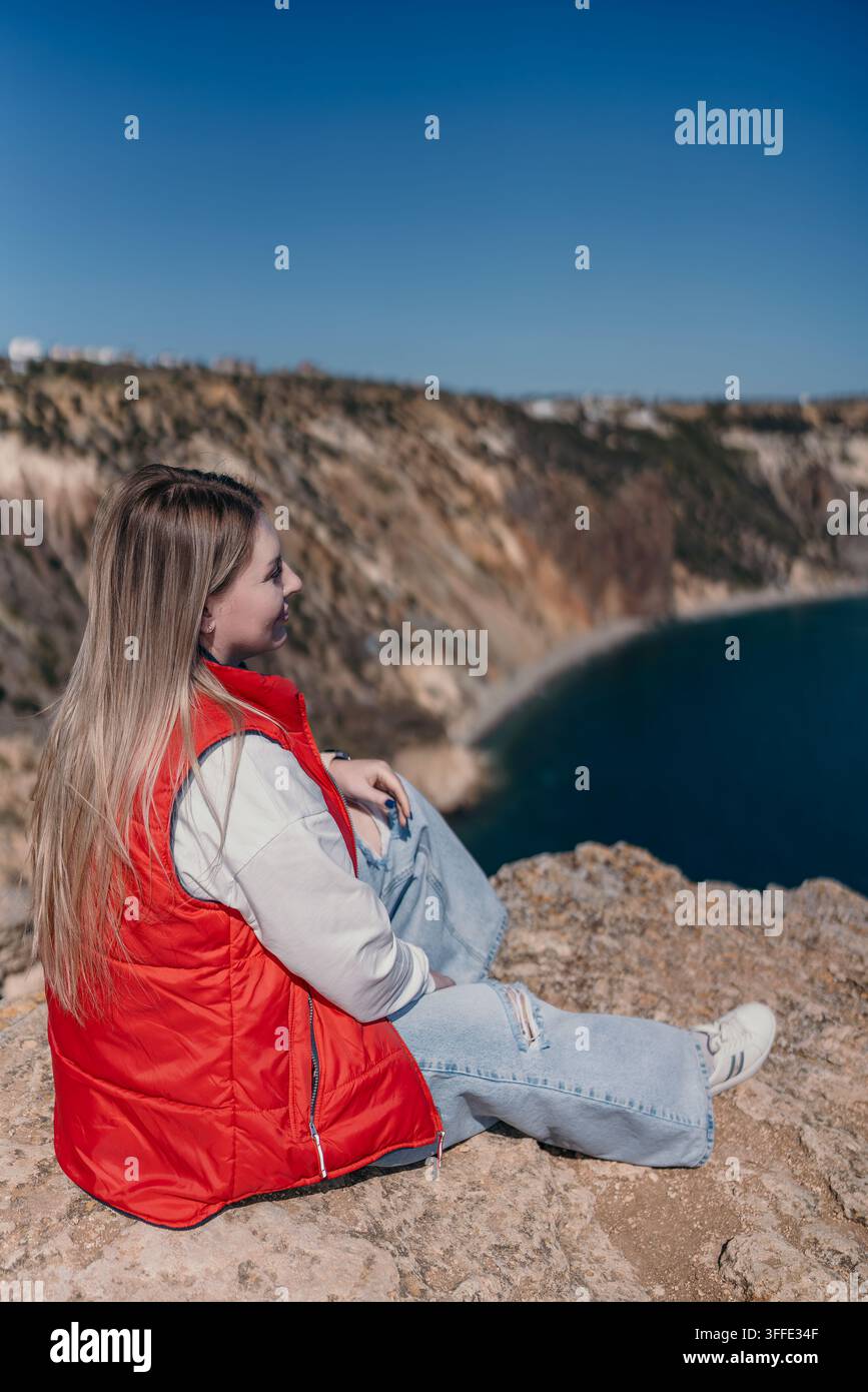 Donna sulla costa a strapiombo seduta su un bordo roccioso che si affaccia su una baia scenografica sotto un cielo blu terso durante un'avventura di viaggio Foto Stock