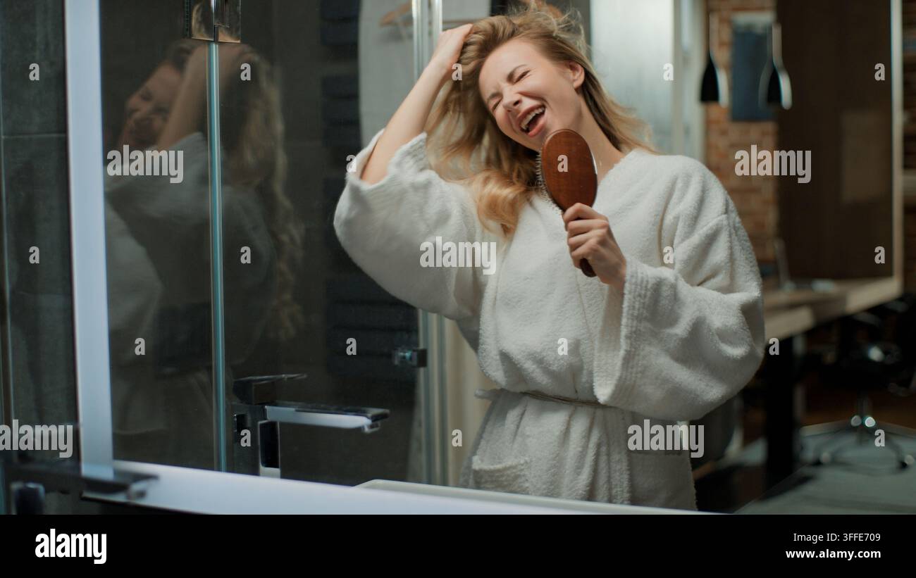 Donna caucasica felice e divertente di mezza età che canta con una spazzola per capelli, toccando capelli ricci morbidi e lucenti, divertendosi, facendo la scema e guardando il riflesso nello specchio a Foto Stock
