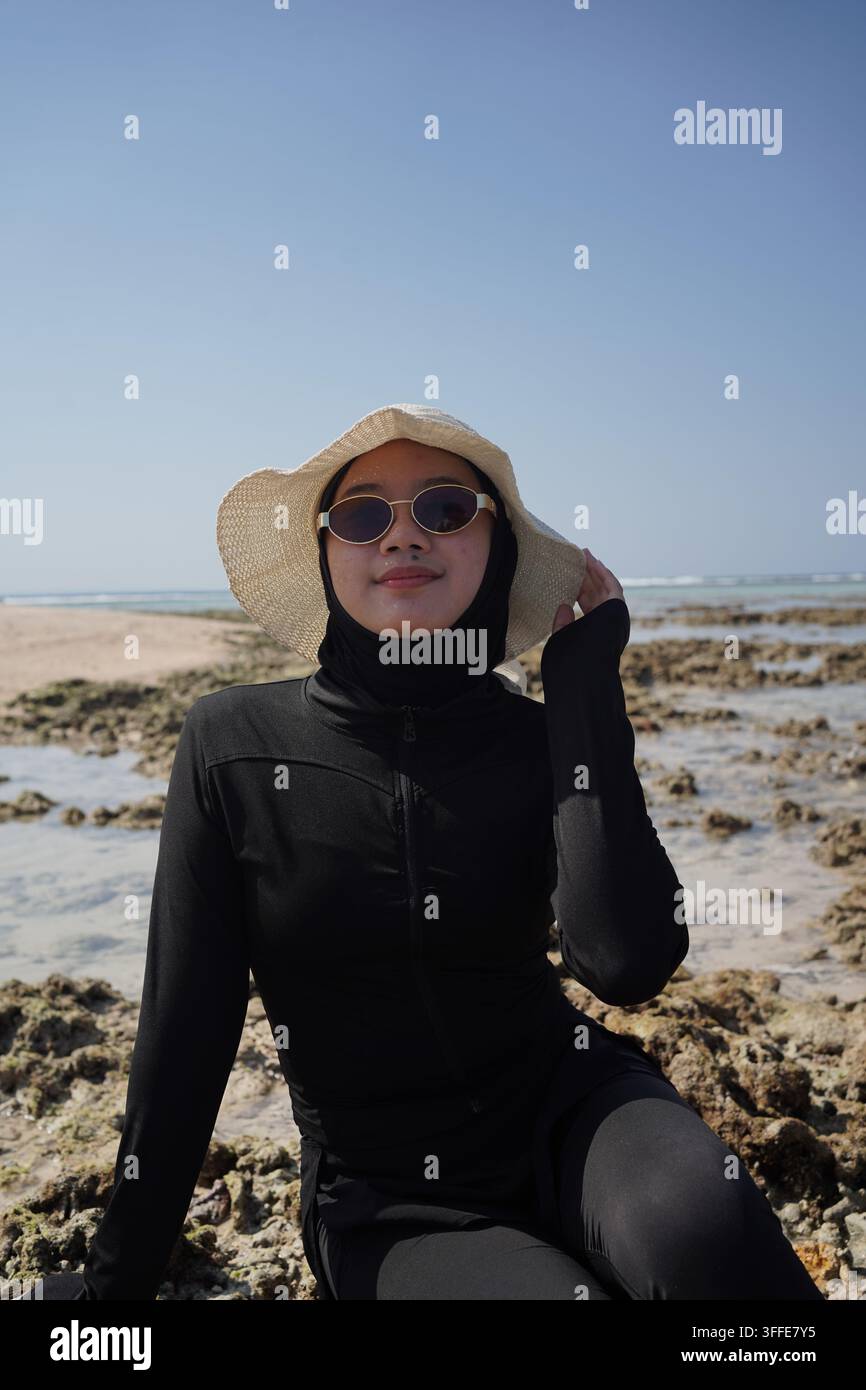 Una donna asiatica in un hijab nero, occhiali da sole e un cappello da sole è seduta rilassata su una spiaggia tropicale rocciosa con formazioni coralline e acqua di mare limpida sotto un bri Foto Stock
