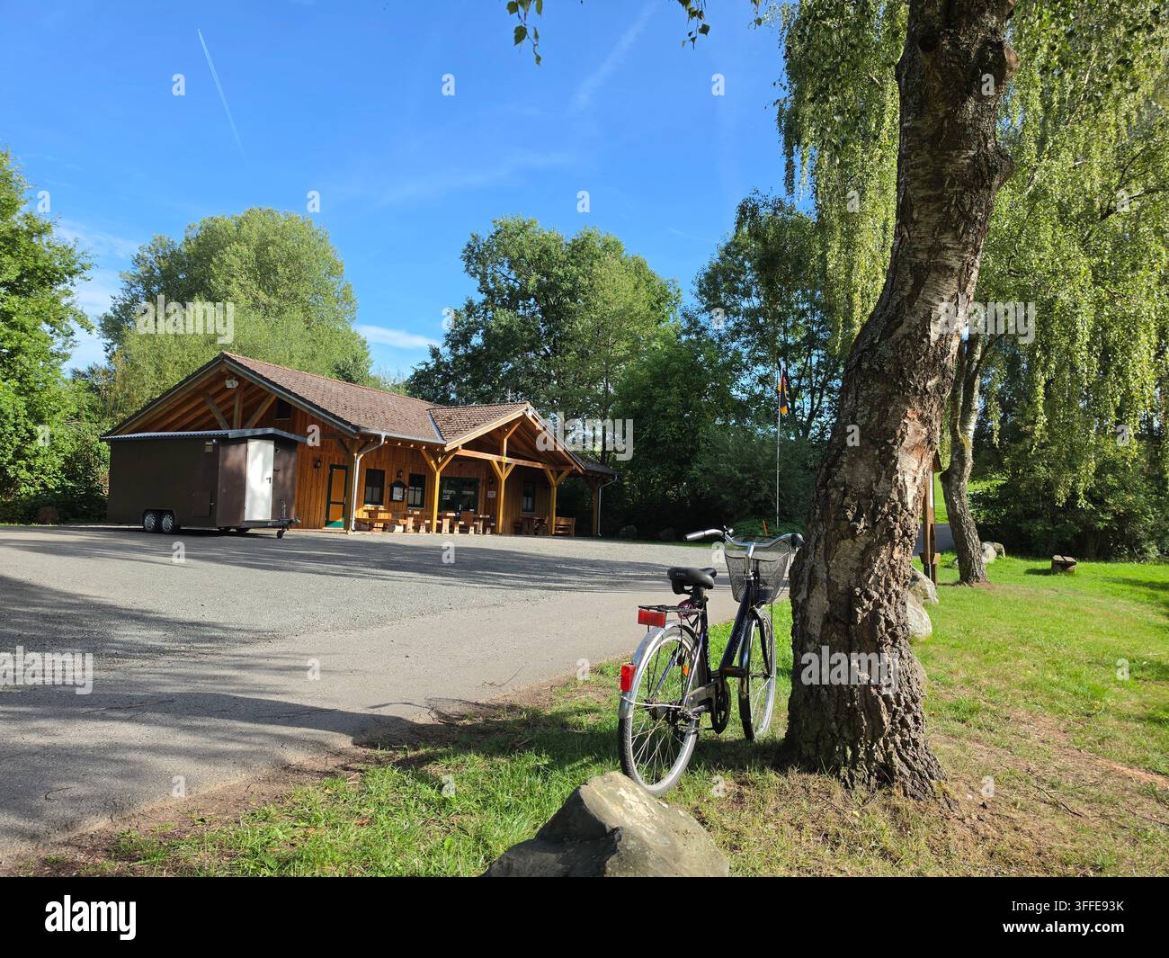Bicicletta parcheggiata vicino a un grosso tronco d’albero vicino a un rifugio in legno in un parco rurale tedesco in una giornata estiva soleggiata con cielo azzurro - Immagine stock catturata con smartphone