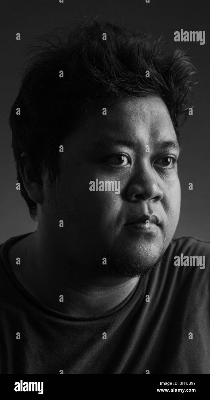 Ritratto in primo piano in bianco e nero di un uomo asiatico adulto che guarda lontano con un'espressione pensierosa su uno sfondo scuro da studio, catturando l'autenticità Foto Stock