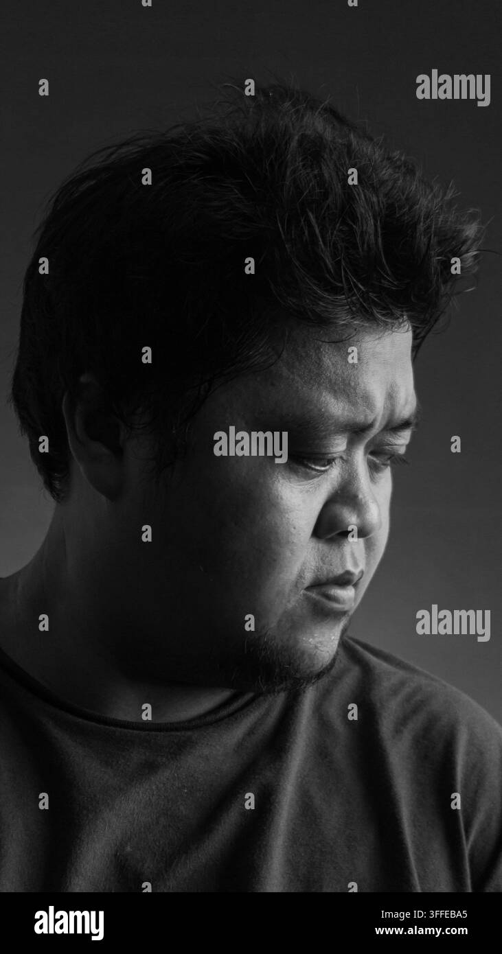 Ritratto in primo piano in bianco e nero di un uomo asiatico adulto che guarda verso il basso con un'espressione triste su uno sfondo scuro da studio, catturando espressioni facciali autentiche Foto Stock