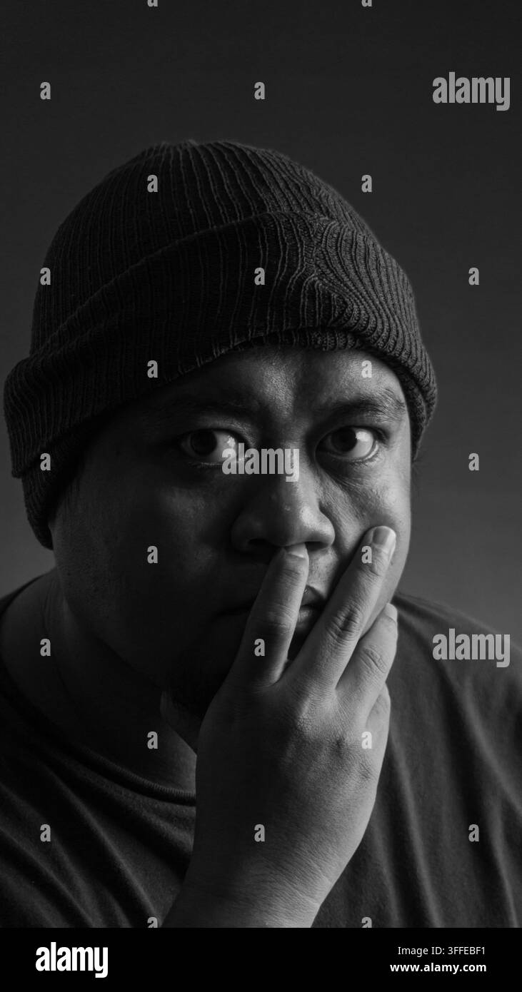 Ritratto in primo piano in bianco e nero di un uomo asiatico adulto che indossa un beanie e con una mano che copre la bocca mentre fissa la fotocamera su uno sfondo scuro Foto Stock