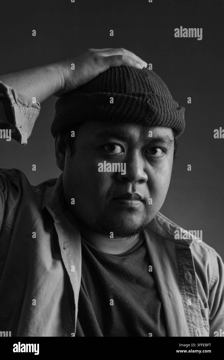 Ritratto in primo piano in bianco e nero di un uomo asiatico adulto con un cappello beanie, una mano appoggiata sulla testa mentre guarda l'obiettivo su uno sfondo scuro Foto Stock