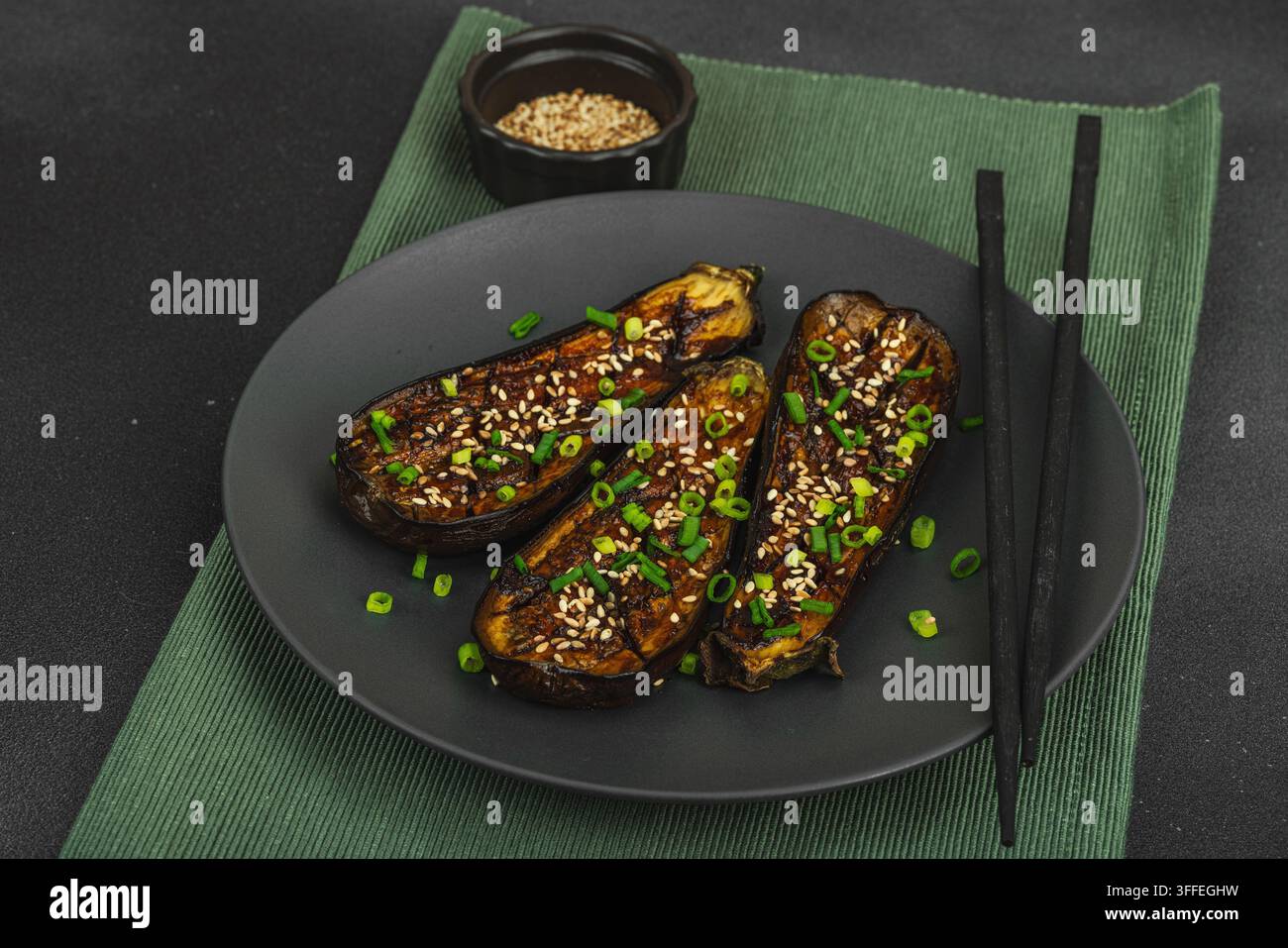 Mezza melanzana glassata al miso con semi di sesamo e cipolla verde su un piatto. Concetto alimentare giapponese di nasu dengaku arrosto con bacchette, cemento di pietra nera Foto Stock