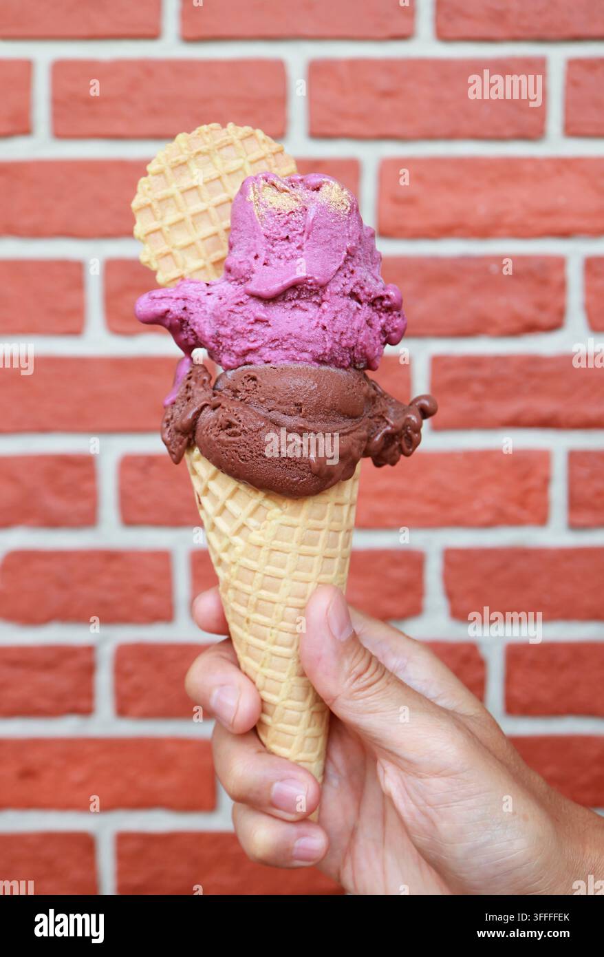 Mano che tiene una doppia porzione di yogurt alla fragola e gelato al cioccolato fondente in un cono contro un muro di mattoni Foto Stock