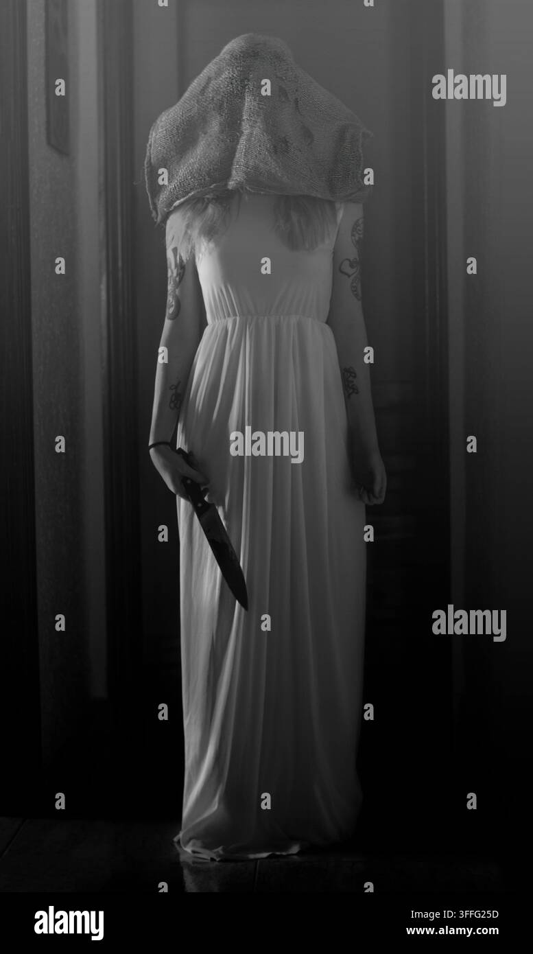 Donna irriconoscibile in un lungo vestito bianco con tessuto strutturato che copre il viso e tiene un coltello affilato Foto Stock