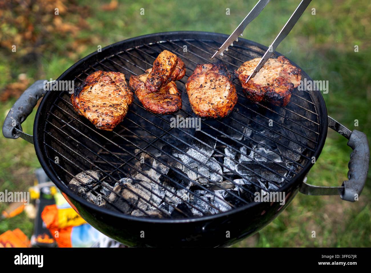 Grigliata di carne stagionata su barbecue a carbone. Pezzi di carne stagionata grigliano su carbone ardente mentre delle pinze di metallo girano un pezzo all'aperto in un verde Foto Stock