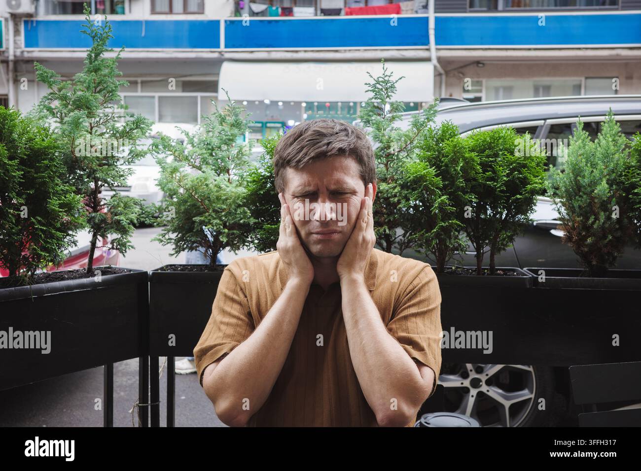 Sovraccarico sensoriale, uomo stressato che preme le mani sulle orecchie con gli occhi chiusi sulla terrazza di un caffè in città, esprimendo stress, sovraccarico, sensibilità al rumore, benessere mentale. Foto Stock