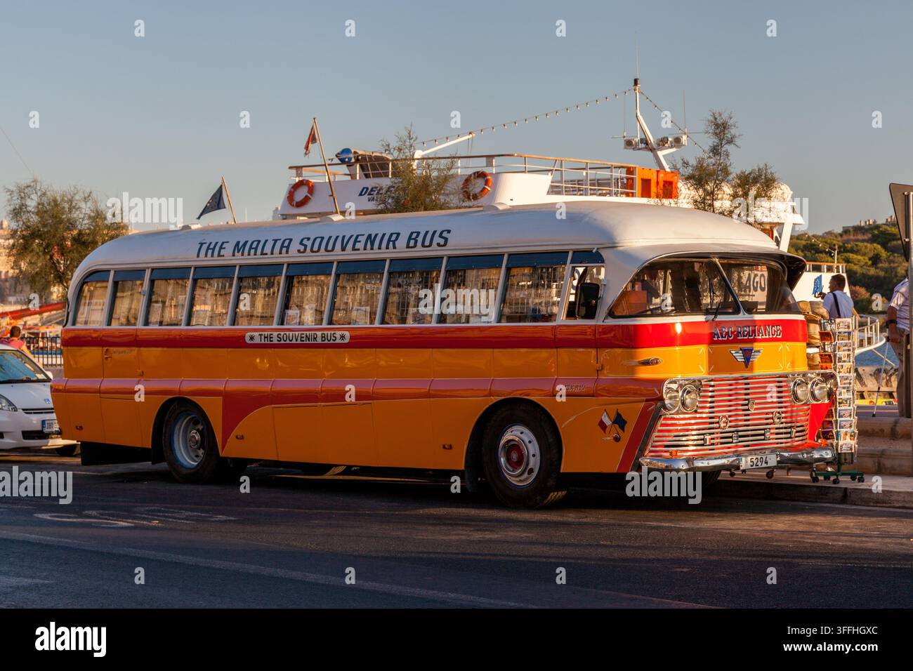 Bus souvenir vintage giallo e rosso di Malta parcheggiato su una strada lungomare, con una barca passeggeri e un espositore di cartoline visibili nelle vicinanze Foto Stock
