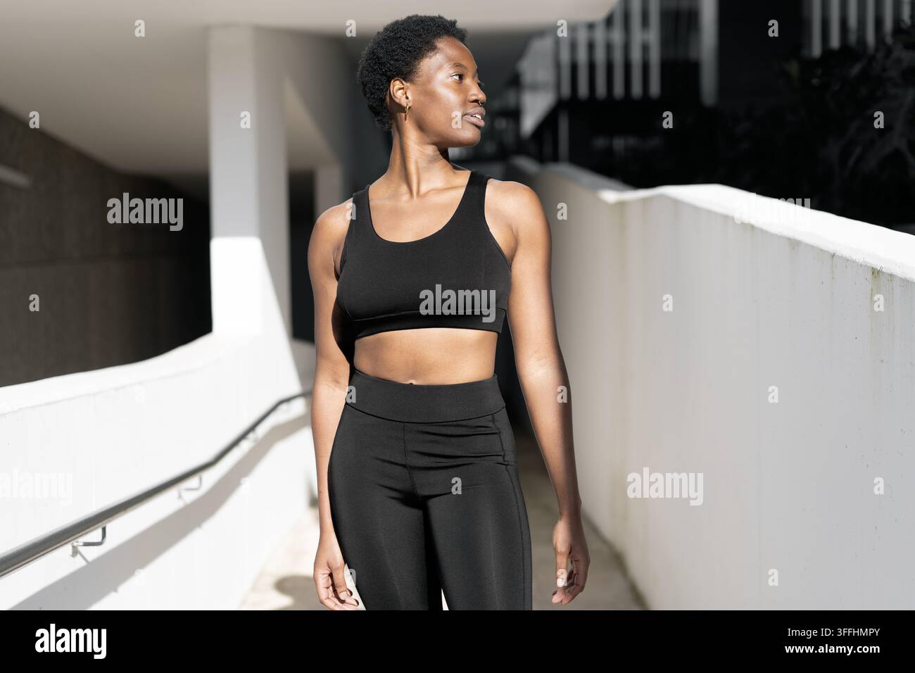 Donna nera in abbigliamento sportivo nero si mostra sicura di sé mentre guarda di lato in uno spazio esterno moderno. Donna afroamericana con capelli naturali corti appare concentrata e pronta per l'allenamento fisico. Foto Stock