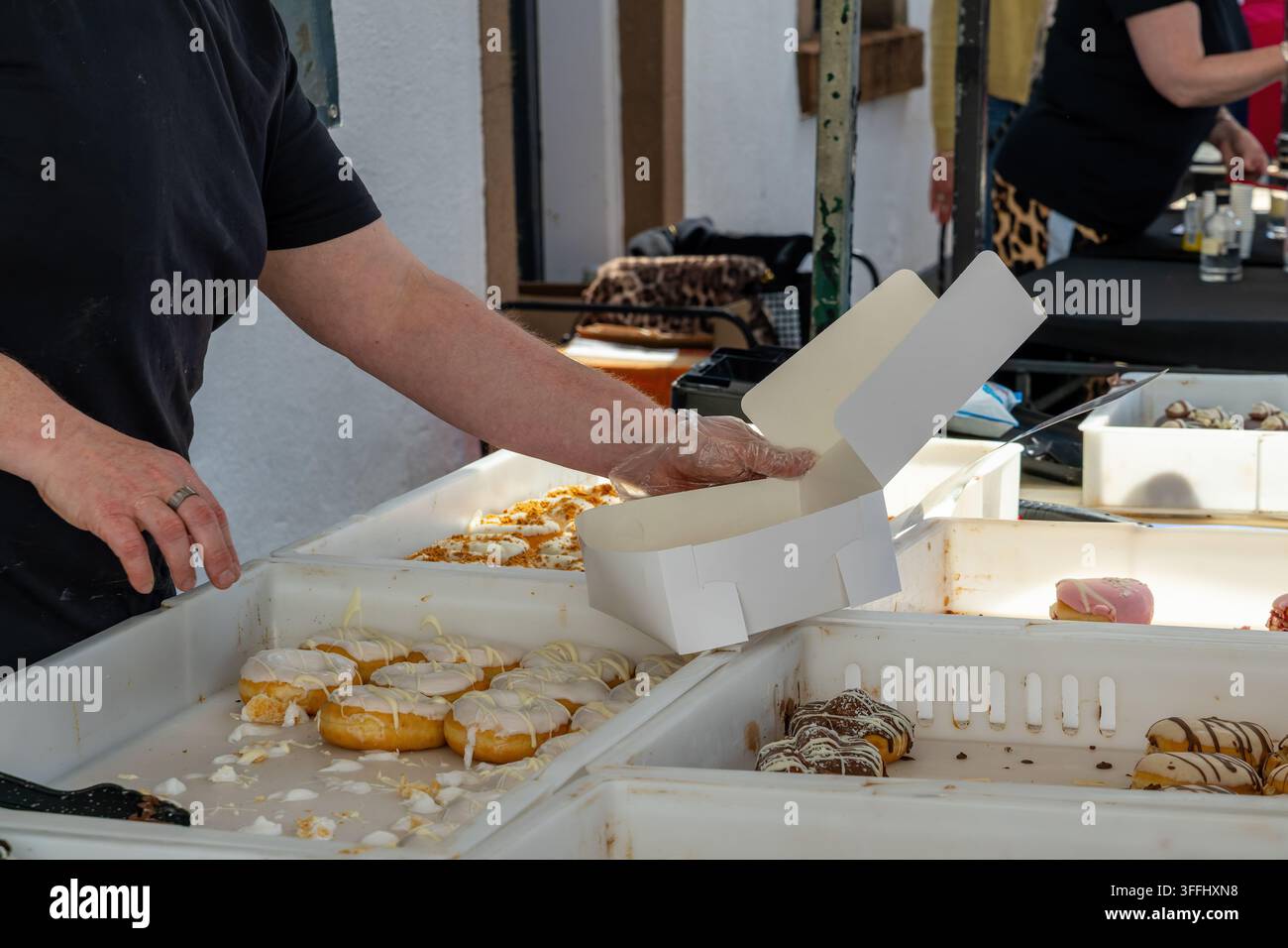 Vendor che indossa guanti monouso posiziona ciambelle decorate in una scatola di cartone bianco in un affollato mercato all'aperto, vassoi che mostrano ciambelle glassate al bianco e al cioccolato Foto Stock