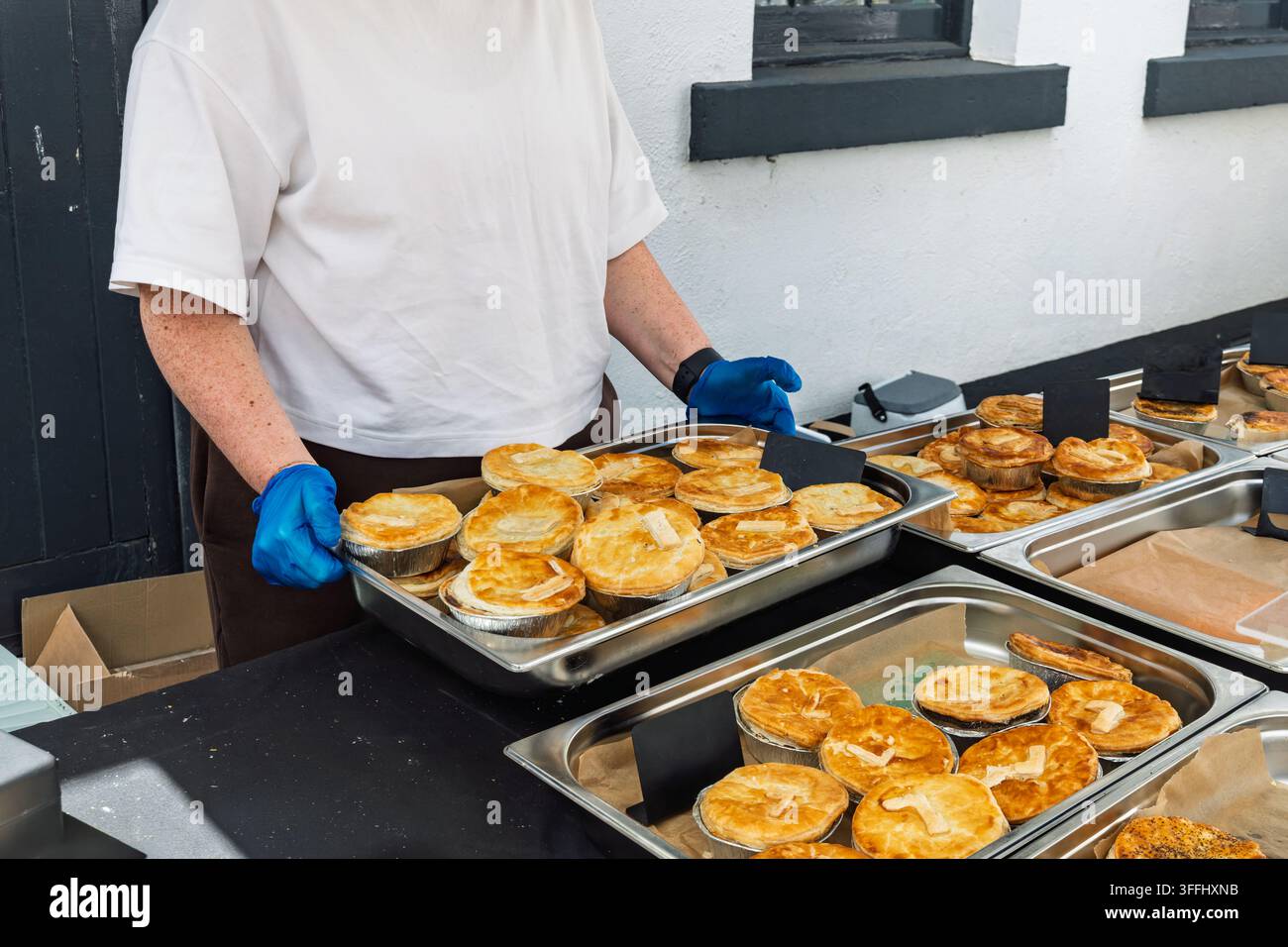 Lavoratore con guanti monouso blu e grembiule marrone maneggia più vassoi di torte di sfoglia dorate porzionate singolarmente in contenitori di alluminio, pronte per Foto Stock