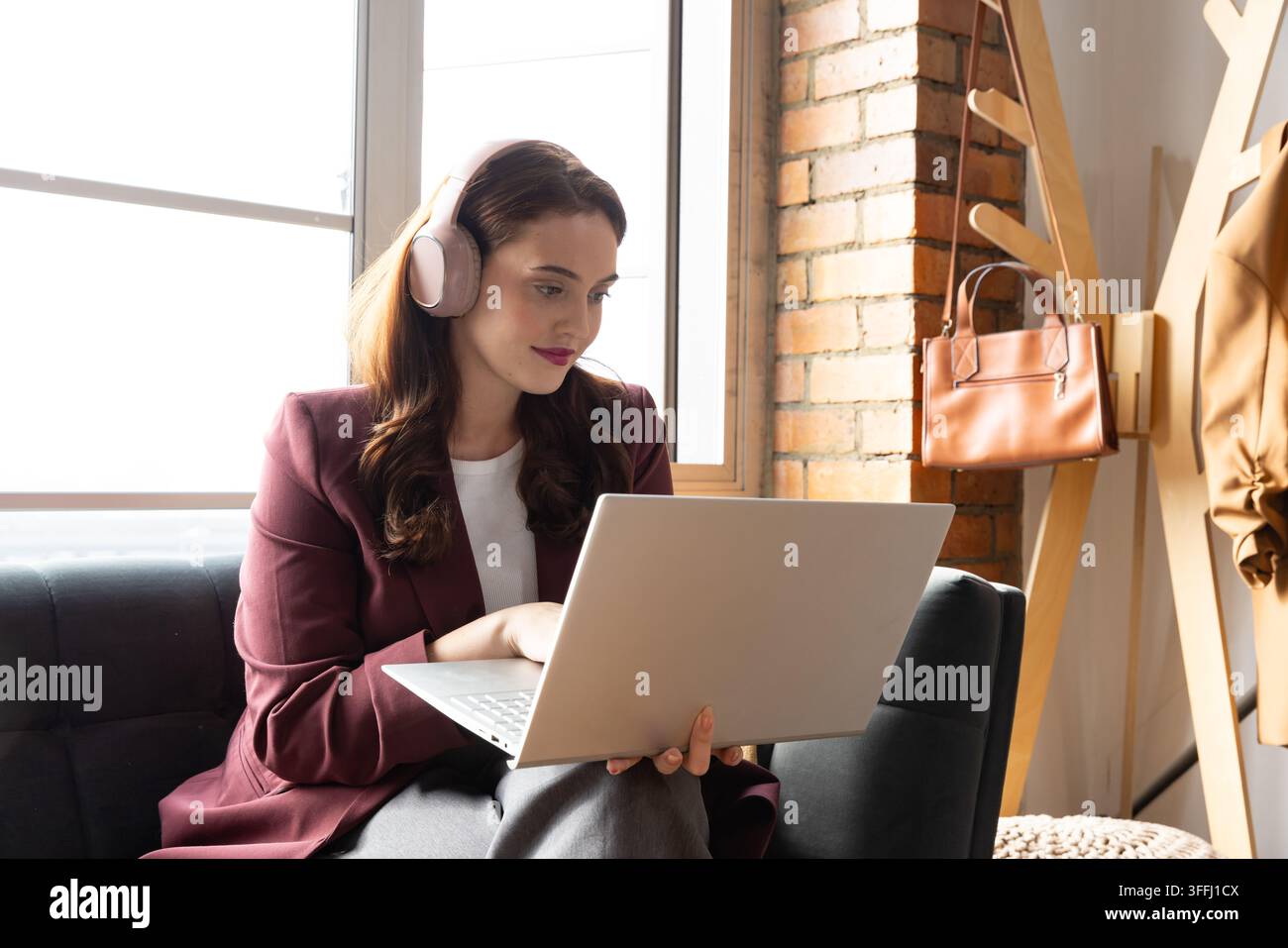 Donna seduta su un divano scuro vicino alla finestra indossa un blazer bordeaux, tiene un laptop e delle cuffie rosa Foto Stock