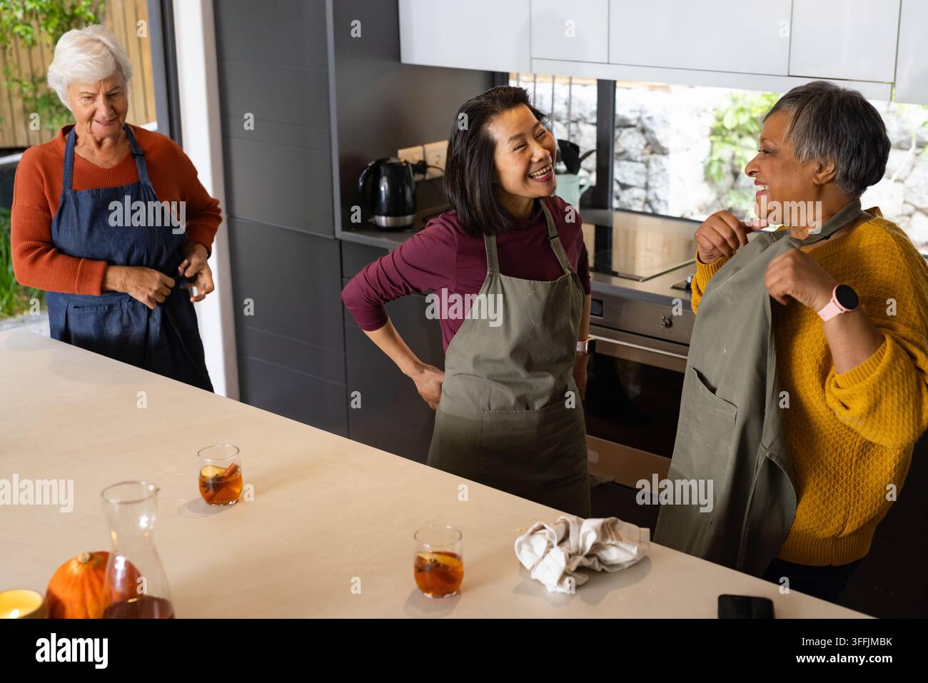 Diverse amiche anziane che ridono indossando grembiuli e preparano bevande ghiacciate vicino a una zucca sull'isola della cucina Foto Stock