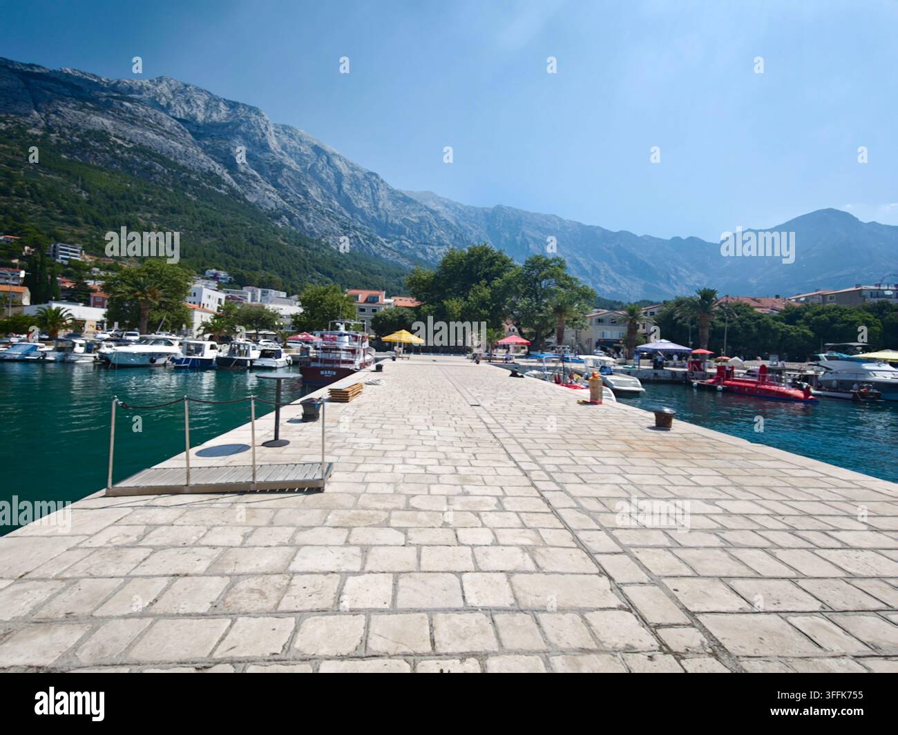Un piano grandangolare di un molo lastricato di pietra che si estende nel mare azzurro con yacht ormeggiati nel porto di Baska Voda. - Immagine stock catturata con smartphone
