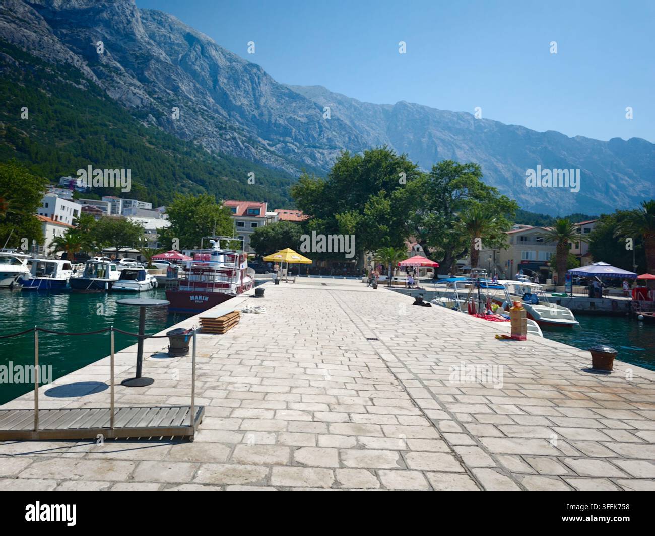 Un piano grandangolare di un molo lastricato di pietra che si estende nel mare azzurro con yacht ormeggiati nel porto di Baska Voda. - Immagine stock catturata con smartphone