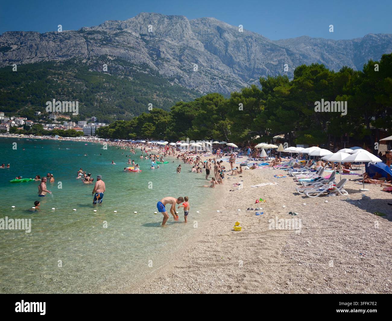 Vibrante scena estiva alla spiaggia di Nikolina, con turisti che si rilassano su rive di ciottoli, nuotano in acque turchesi limpide, sotto un imponente picco montuoso - Immagine stock catturata con smartphone