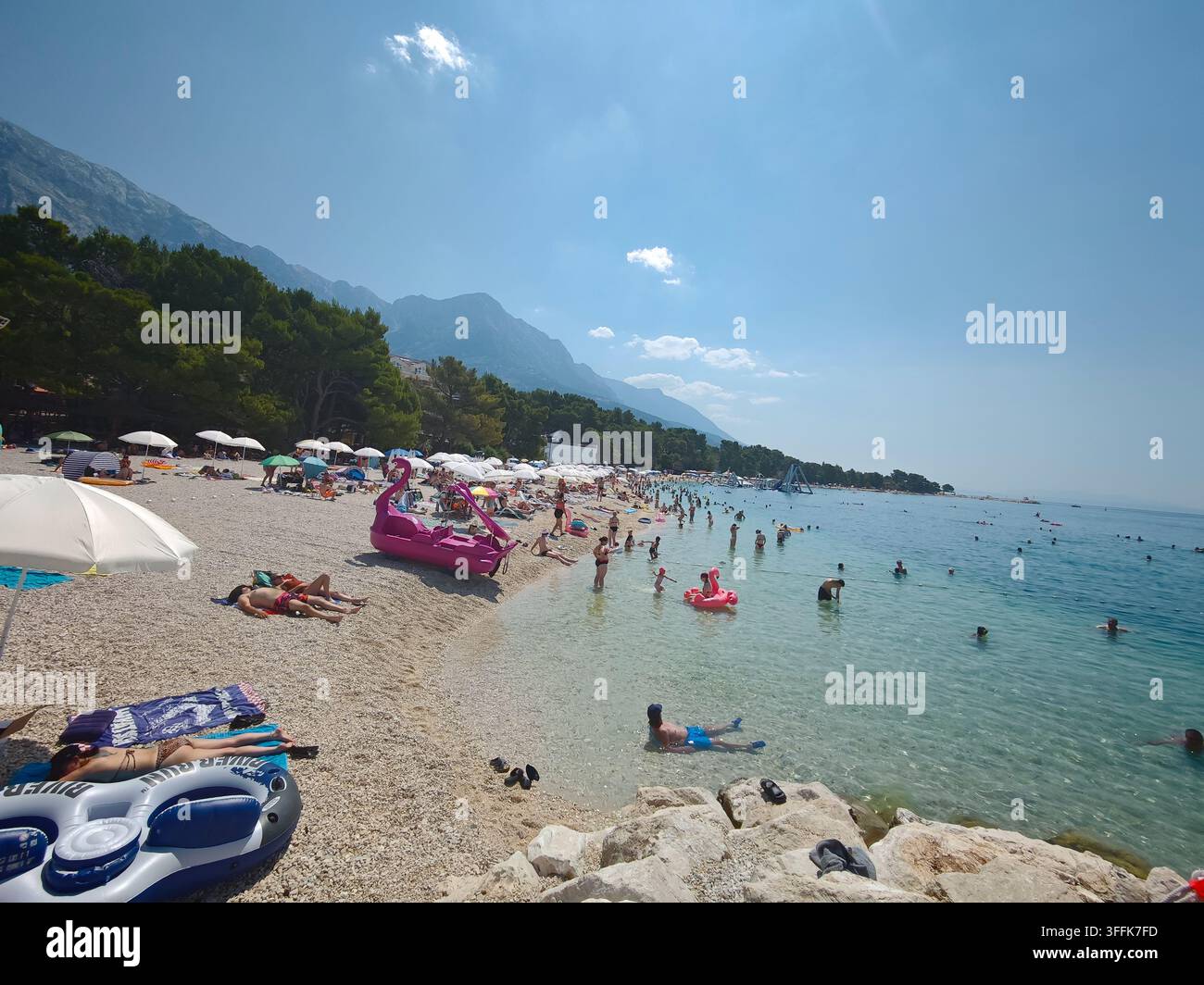 Vibrante scena estiva alla spiaggia di Nikolina, con turisti che si rilassano su rive di ciottoli, nuotano in acque turchesi limpide, sotto un imponente picco montuoso - Immagine stock catturata con smartphone