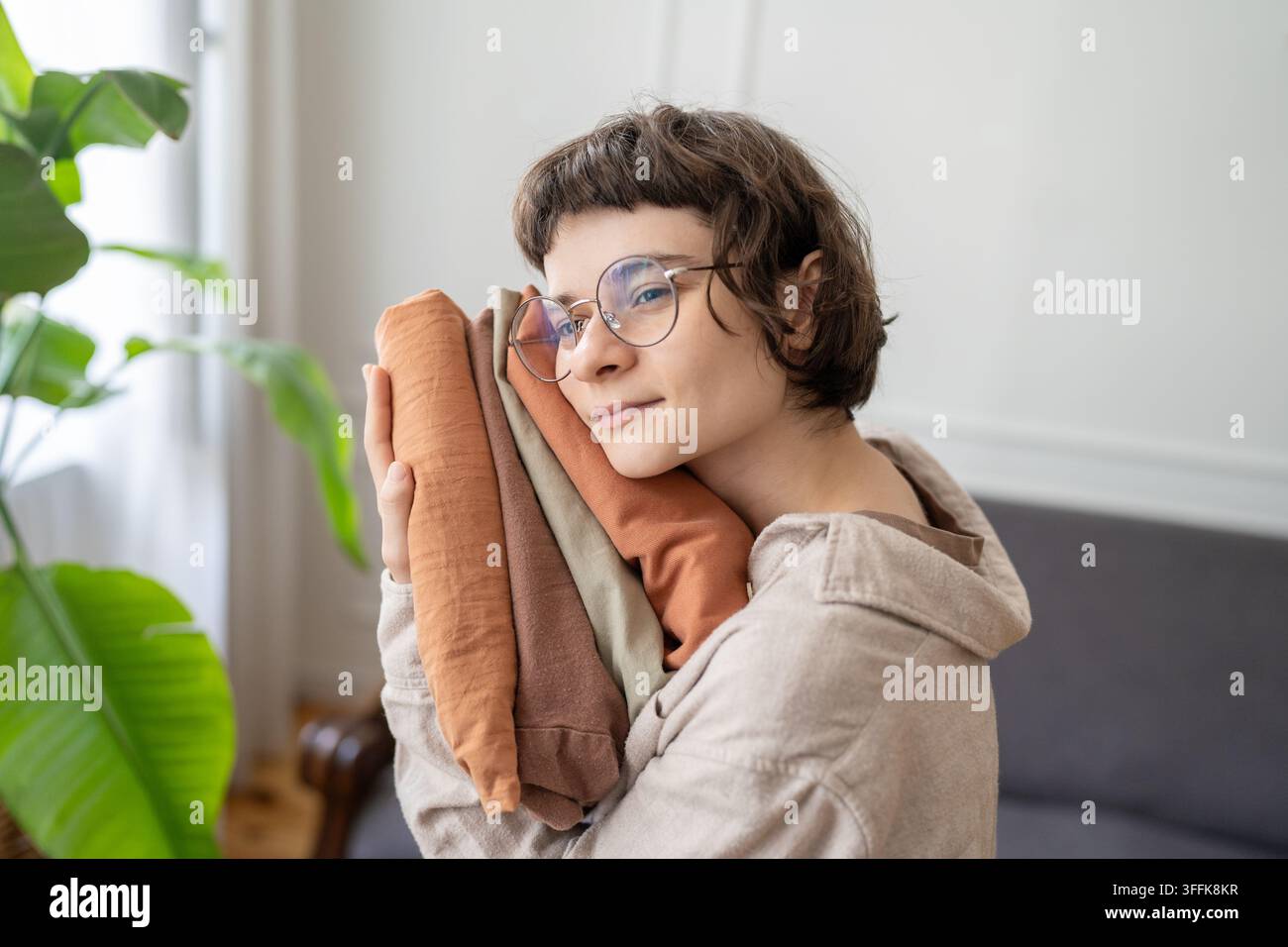 Donna calma che tiene vestiti di lino naturale piegati mentre sceglie articoli di guardaroba sostenibili a casa. Foto Stock