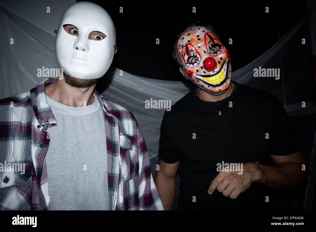 Due uomini adulti che posano con maschere spaventose di Halloween su uno sfondo scuro decorato durante una festa in costume. Foto Stock